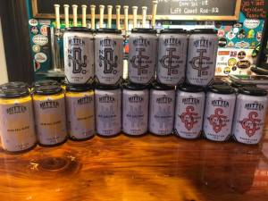 Twelve canned beers stacked on a wooden bar in front of beer taps.