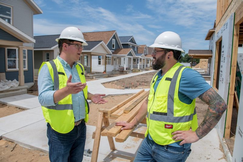 Stephen Wooden discussing a housing construction project with a former colleague. Both are wearing yellow vests and hard hates, standing by pile of lumber in front of houses.