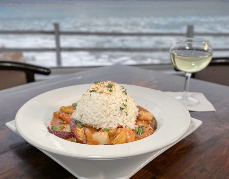 a plate of food with a glass of white wine on a table in front of a view of the waterfront